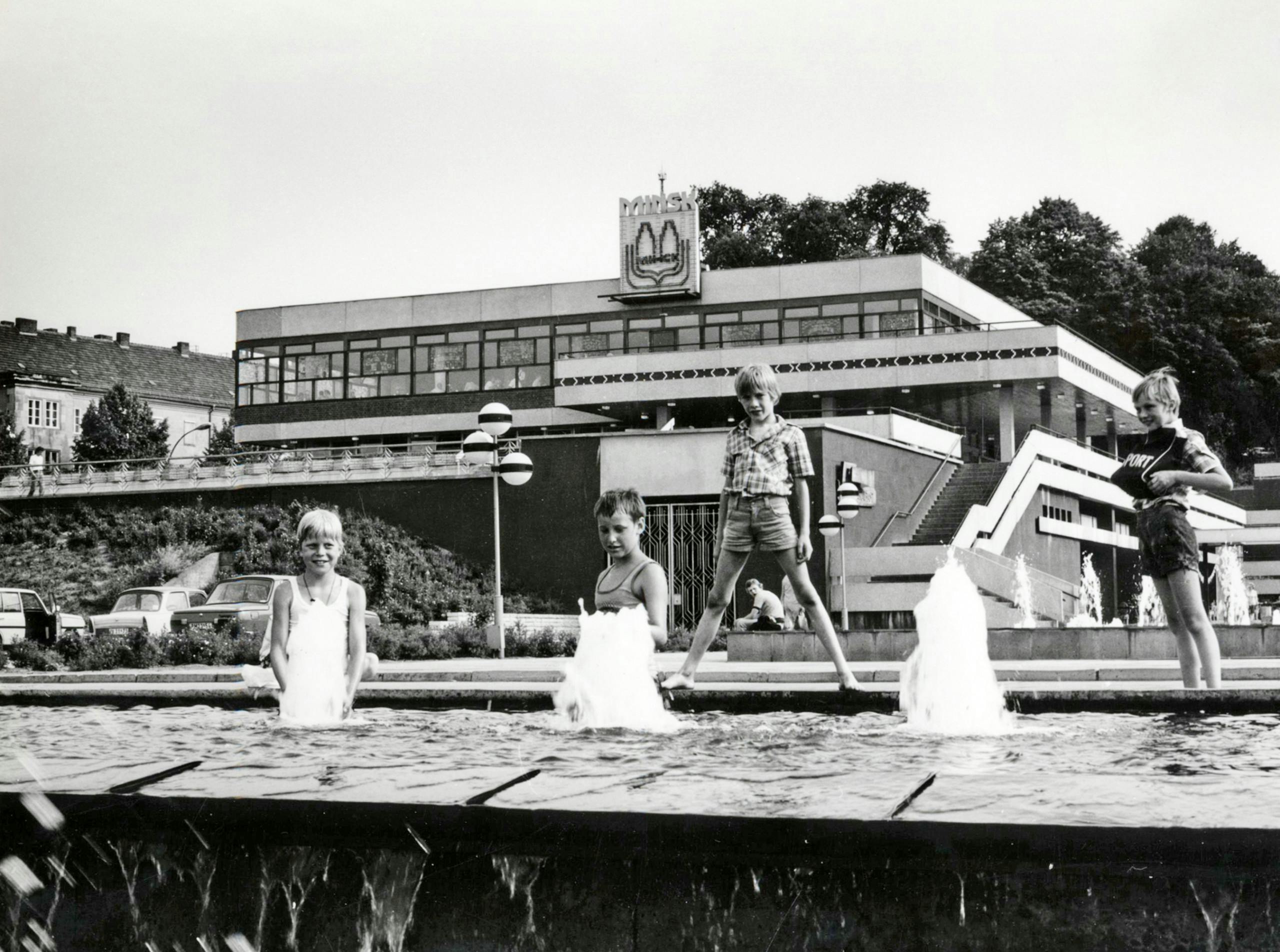 Black and white photo: in the foreground, four children dressed in summer clothes are playing by a fountain. A boy stands on the edge of the fountain with his legs apart. In the background there is the former terrace restaurant Minsk.