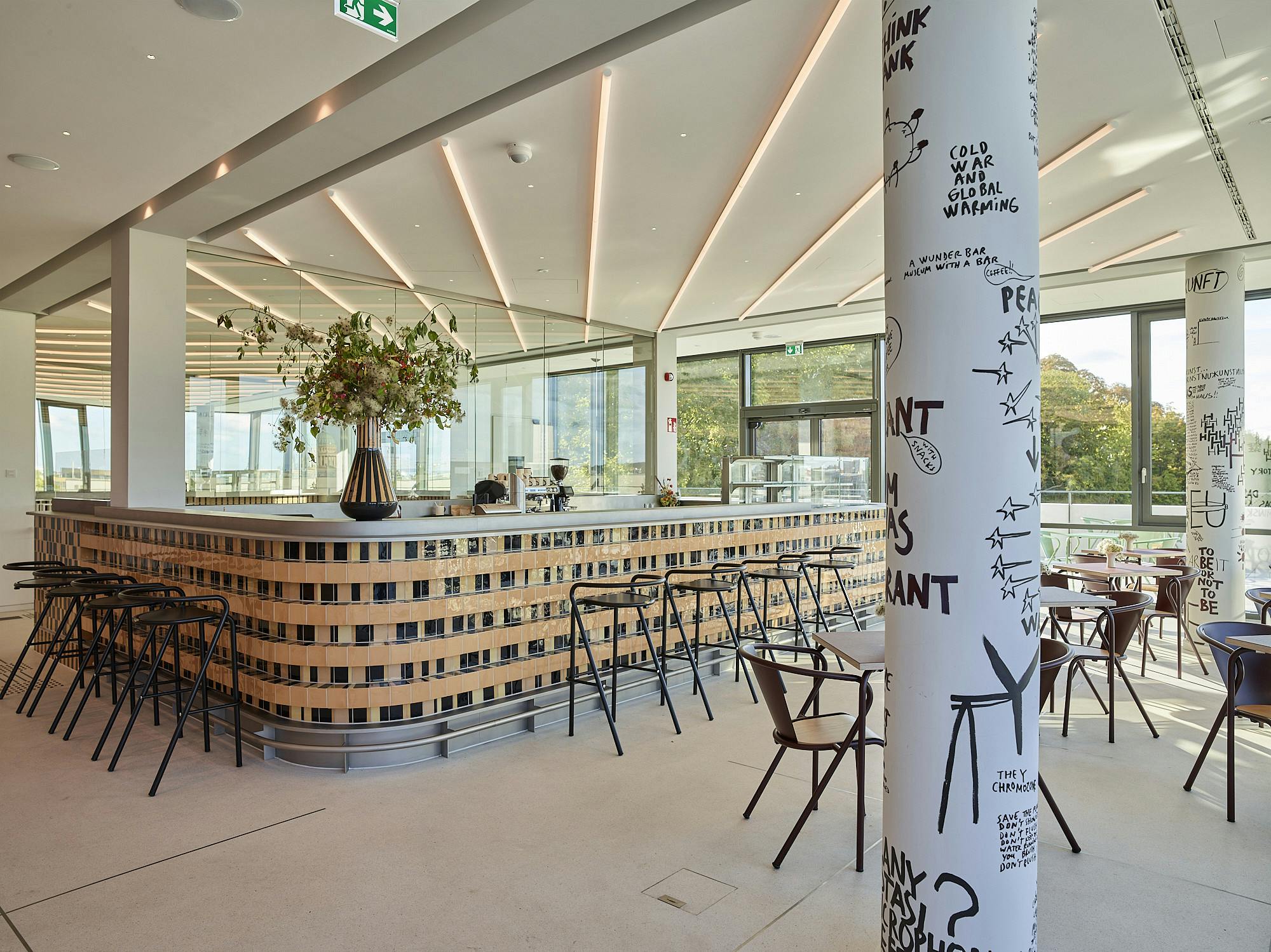 Photo: View into Café Hedwig at DAS MINSK. Behind a column painted with black words and signs by the artist Dan Perjovschi there is a bar counter decorated with brown and black tiles.
