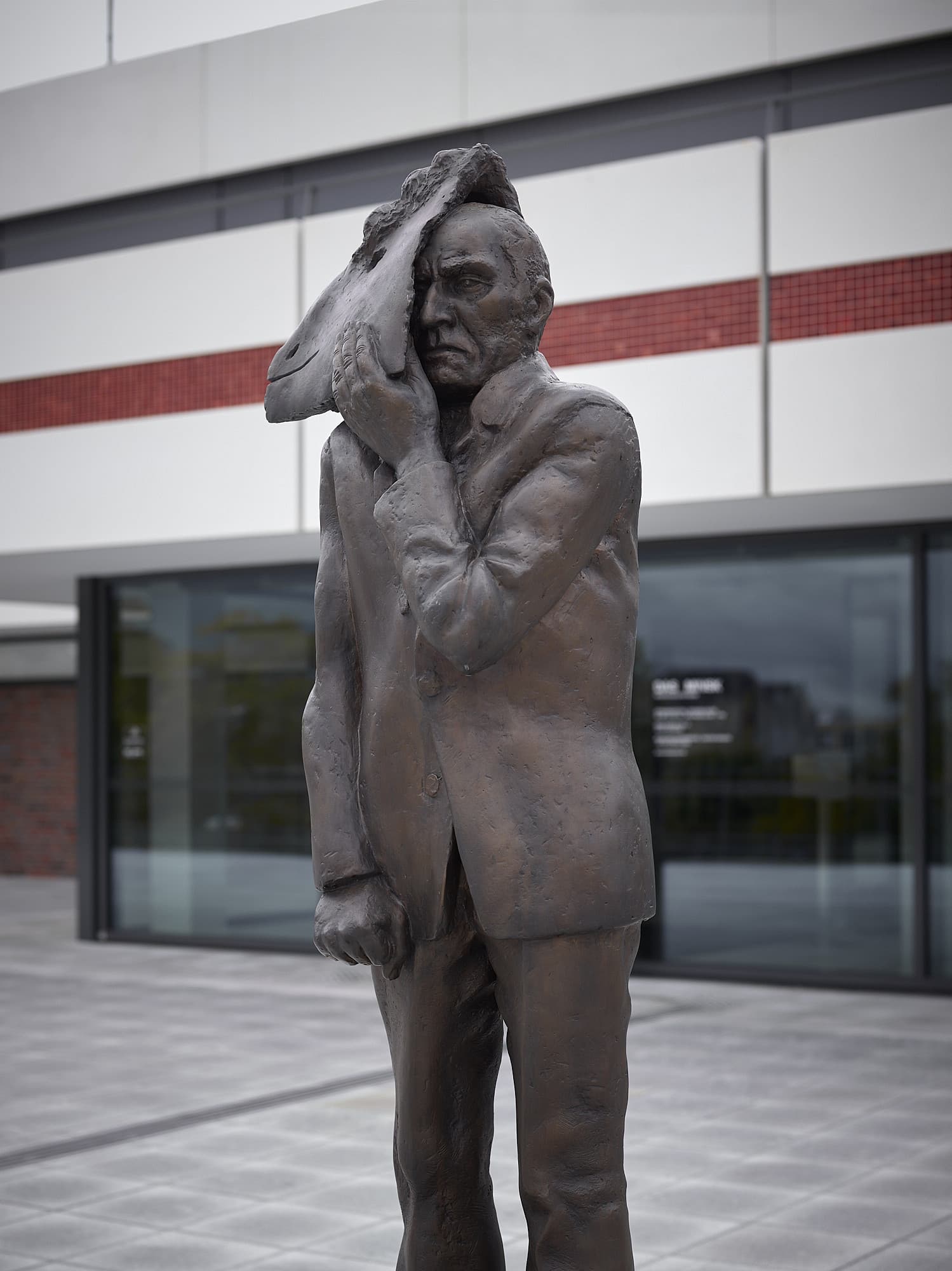 Photo: A life-size bronze sculpture stands on the terrace in front of DAS MINSK. The upright man in a suit is holding a sheep mask with one hand halfway up his serious face.