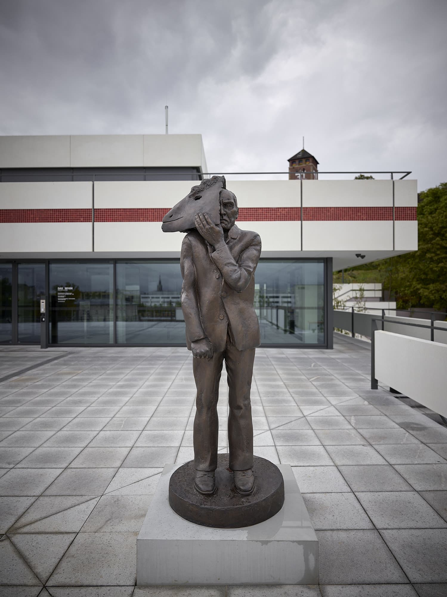 Foto: auf der Terrasse vor dem MINSK steht eine Bronzeskulptur in Lebensgröße. Der aufrechtstehende Mann im Anzug hält mit einer Hand eine Schafsmaske halb vom ernsten Gesicht.