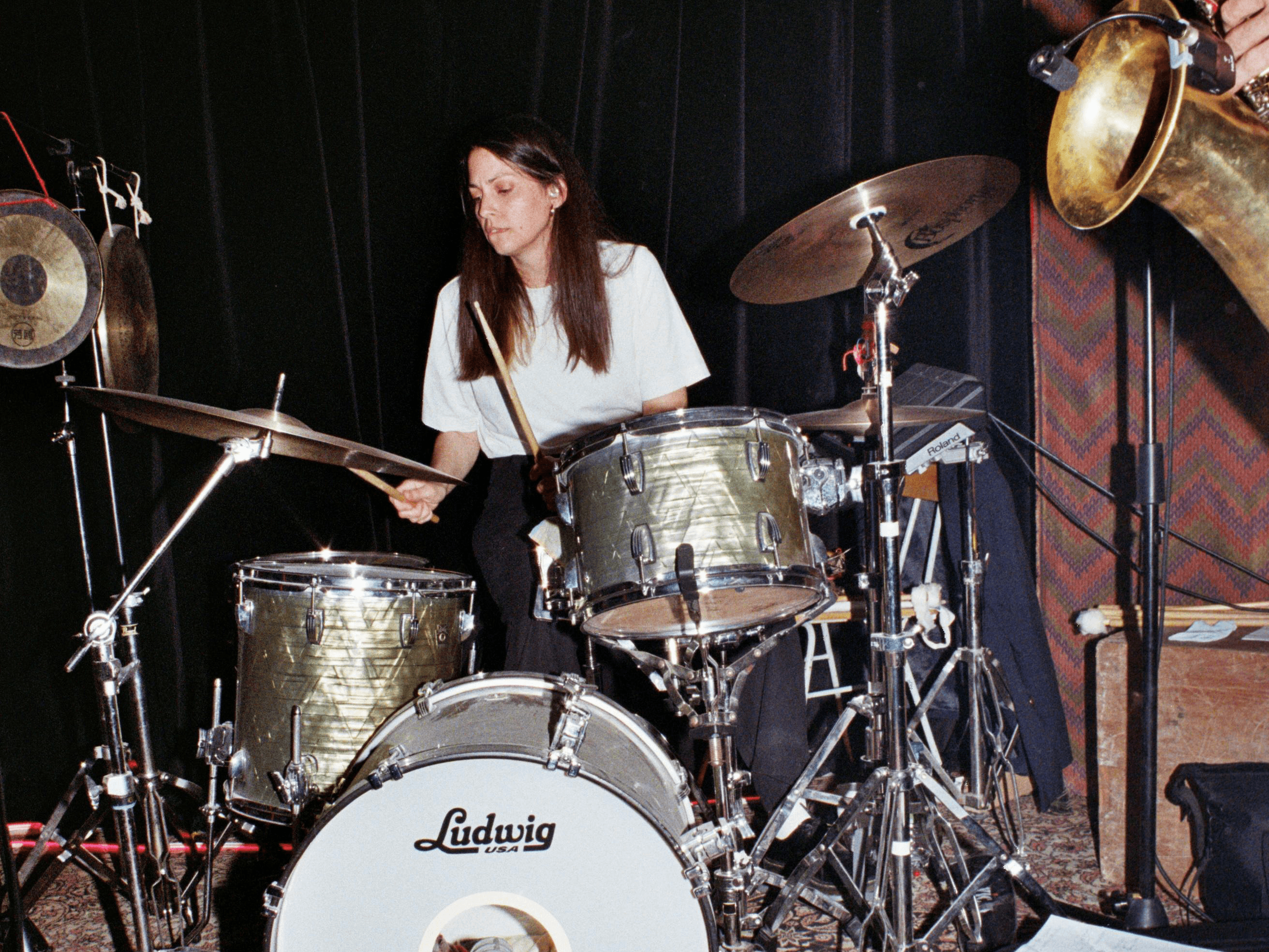 Foto: Die Musikerin Danae Palaka spielt auf der Bühne Schlagzeug, im Vordergrund ist ein Saxophon angeschnitten. 