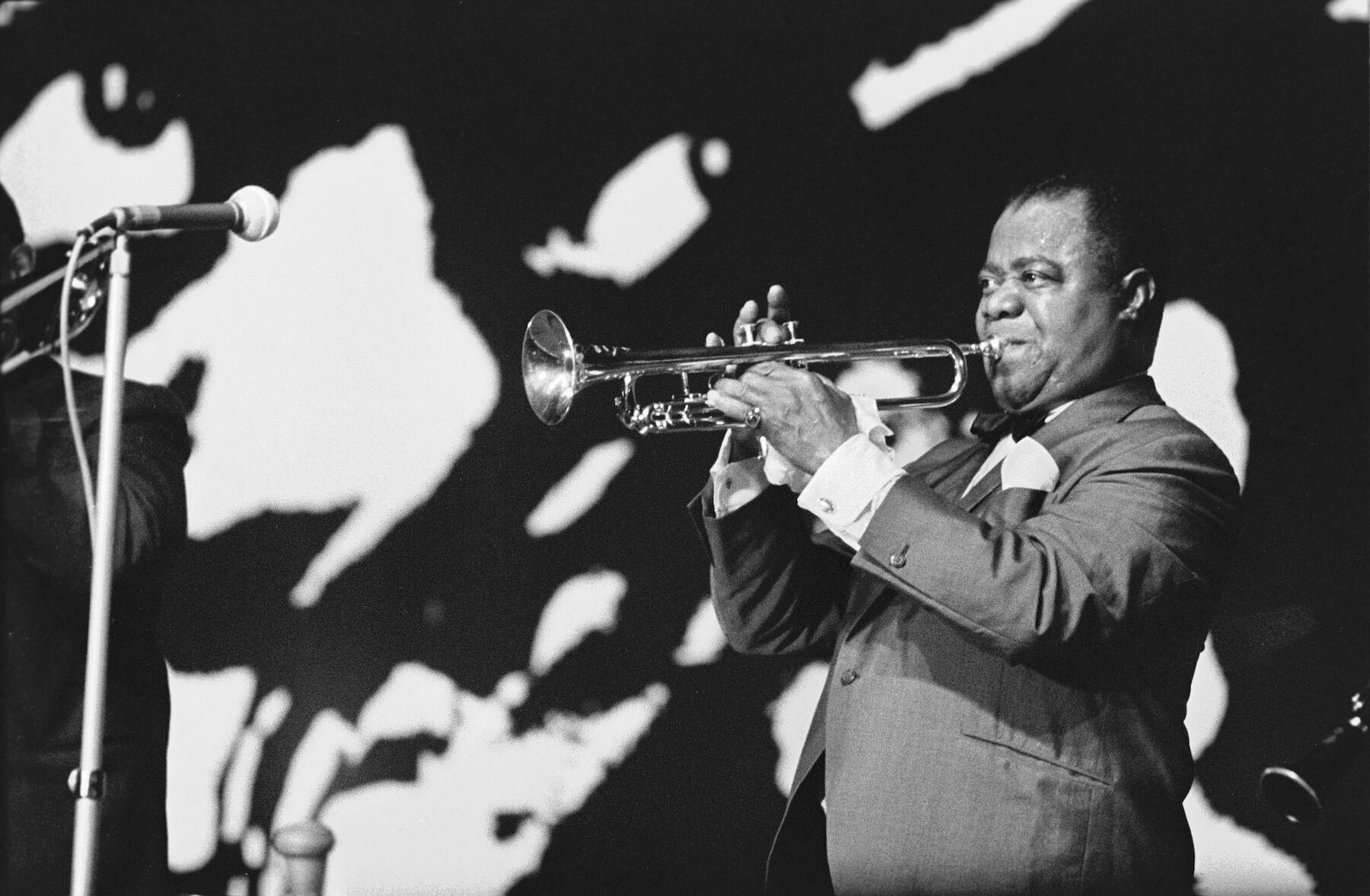 Black and white photo: Louis Armstrong stands on a stage playing the trumpet. Other musicians are only vaguely indicated in front of the black background.