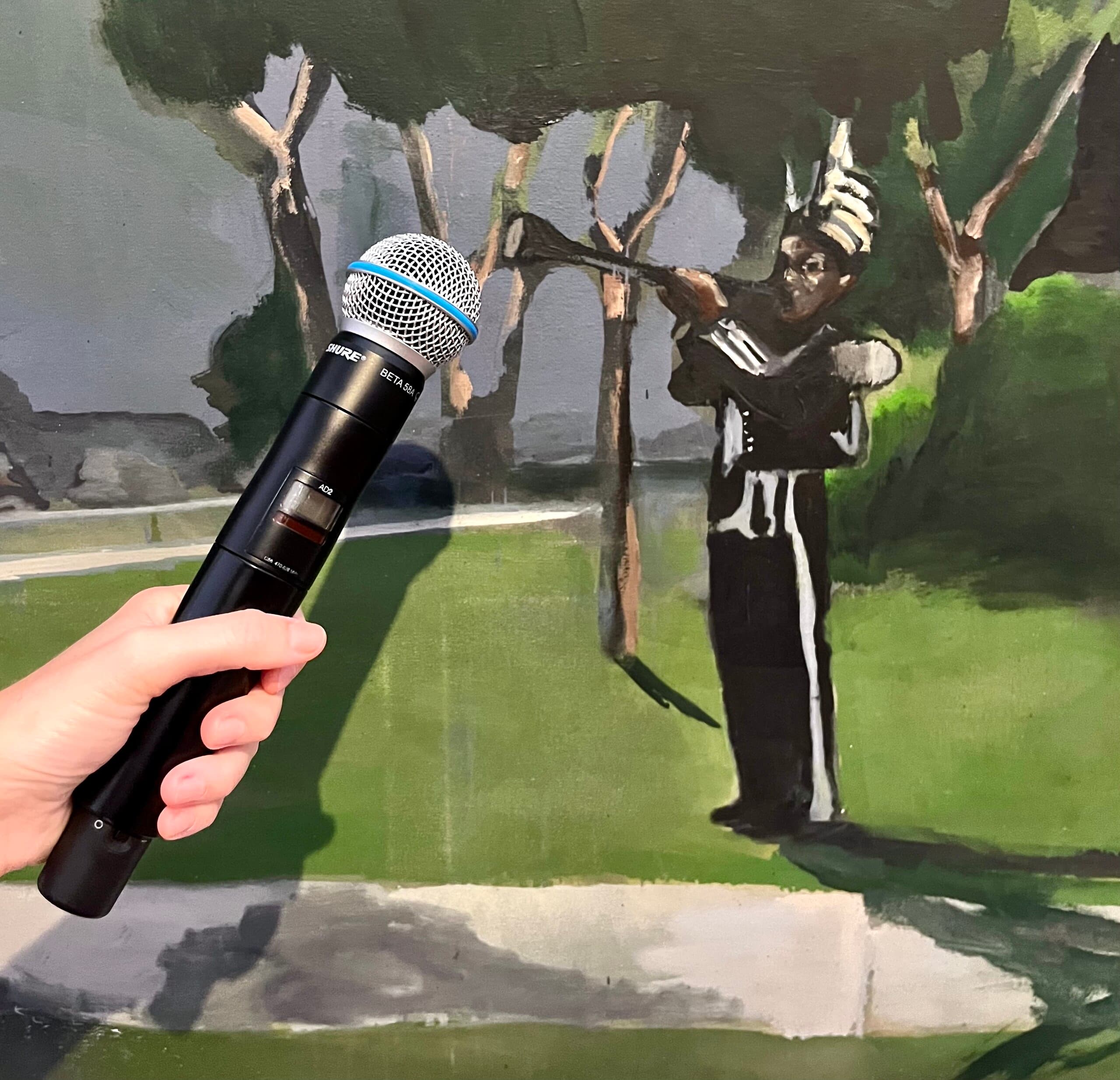 Photo: A hand holds a microphone in front of an artwork showing a Black person playing a trumpet. It illustrates the possibilities of a sound workshop.  