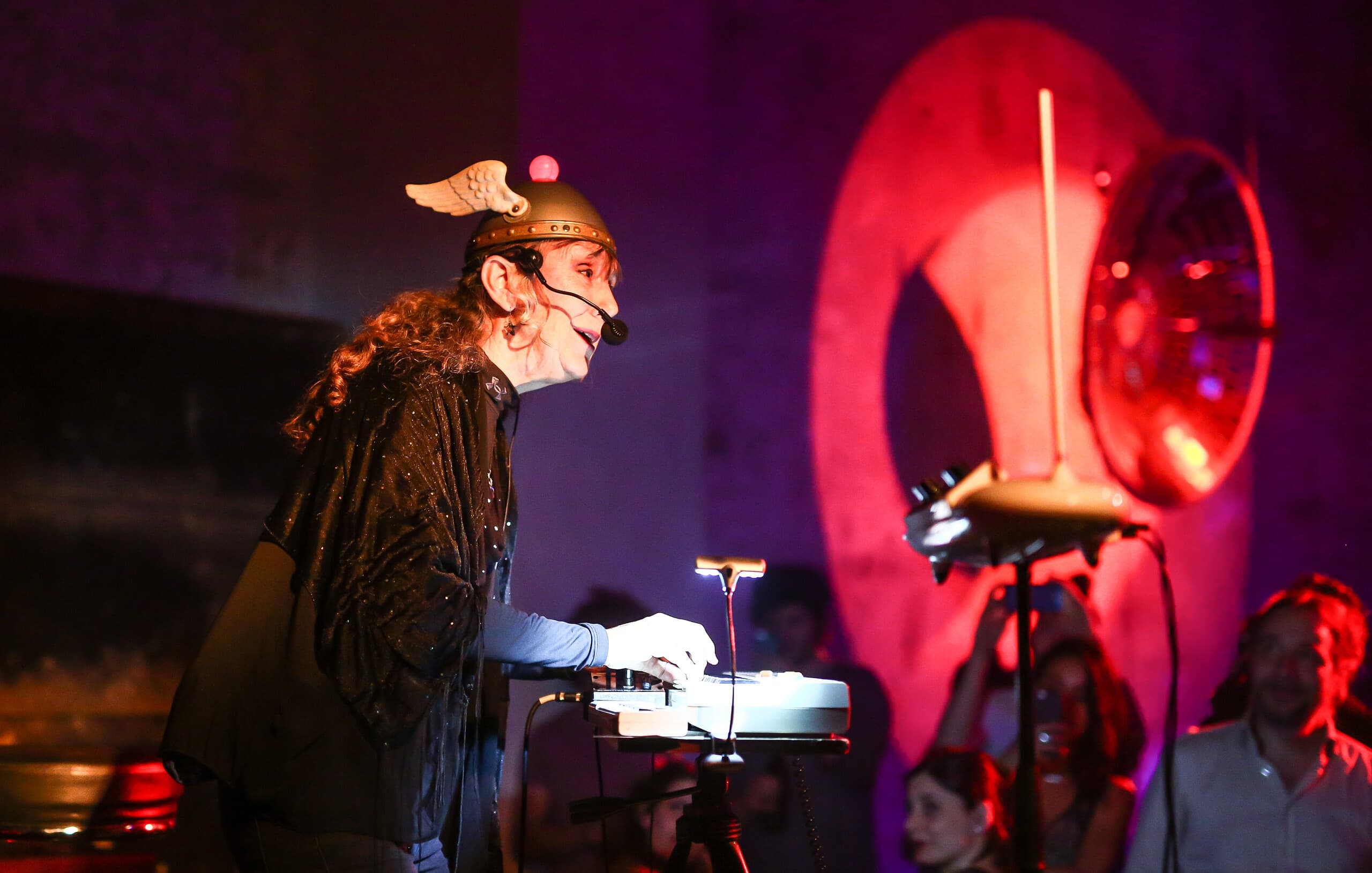 Photo: Musician The Space Lady plays the keyboard on stage and sings into a headset microphone. She wears a helmet with wings. 