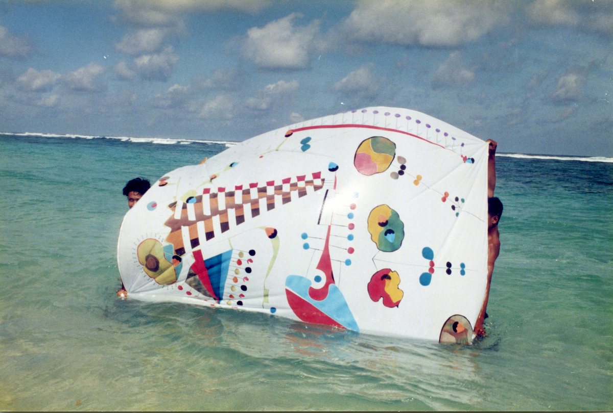 Photo: Two people are standing in shallow seawater, stretching a large bright sheet of fabric with colorful, varied shapes between them. Above them is a blue sky with clouds.